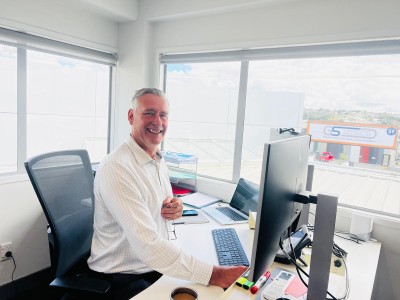 A smiling middle aged man sitting at a computer next to a window