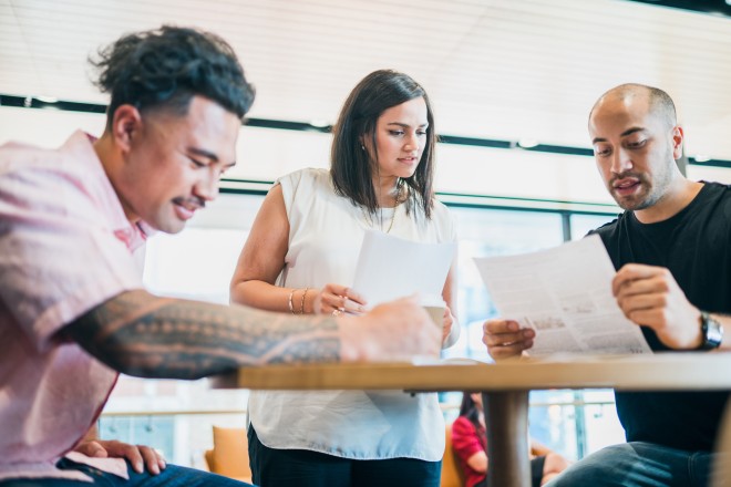 Team of two men and a woman discussing the new strategy. The man in the foreground has a large Pasifika tribal tattoo on his forearm.